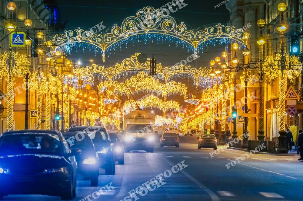 На фотографии запечатлен Невский проспект, украшенный яркой новогодней иллюминацией. Светящиеся арки и гирлянды тянутся вдоль всей улицы, создавая волшебную атмосферу. Автомобили и освещенные фасады зданий дополняют праздничный городской пейзаж. Невский проспект в эту пору превращается в сердце праздничного Санкт-Петербурга, привлекая горожан и туристов.